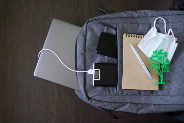 istock Gray textile city backpack, which contains a silver laptop, power bank, smartphone, protective medical mask and a notebook on a wooden background. Study, travel and trip with gadgets 1365651005