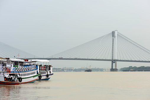 View down river Hooghly flanked by moored boats and Vidyasagar bridge on horizon under morning sky in Kolkata, West Bengal, India.
