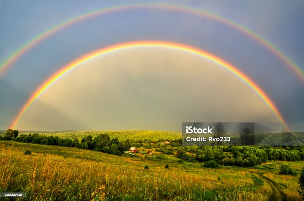 Full arc of double rainbow over summer evening rural landscape - panoramic view Full arc of double rainbow over summer evening rural landscape - panoramic view of hills with deep forests covered, golden colored meadow with sun beams illuminated, village near forest Rainbow Stock Photo
