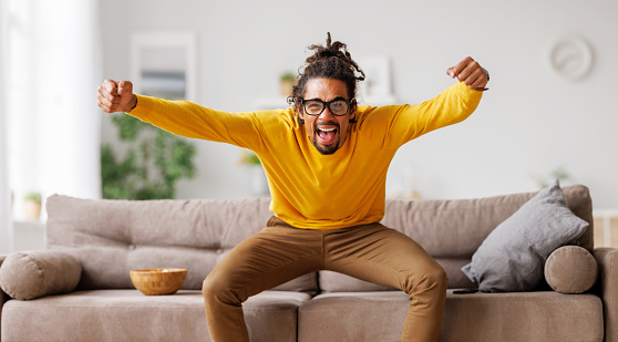 Overjoyed excited young african american man raising arms and celebrating goal while watching football match on tv at home during championship, selective focus. Sport, people and hobby concept
