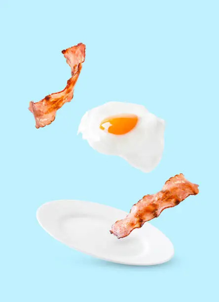 Bacon and egg as English breakfast levitate over a plate on a blue background.