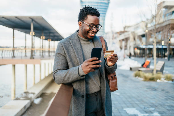 Afro american businessman using smart phone on city street Middle age Afro American businessman standing on street and using smart phone and bank credit card for online payment. businessman african descent on the phone business person stock pictures, royalty-free photos & images