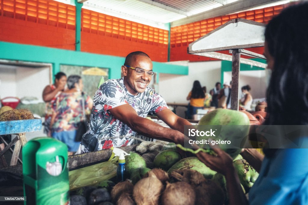 smiling young market vendor giving fresh coconut to female client smiling young market vendor giving fresh coconut to female client at market stall at public market in Bahia, Brazil Agricultural Fair Stock Photo