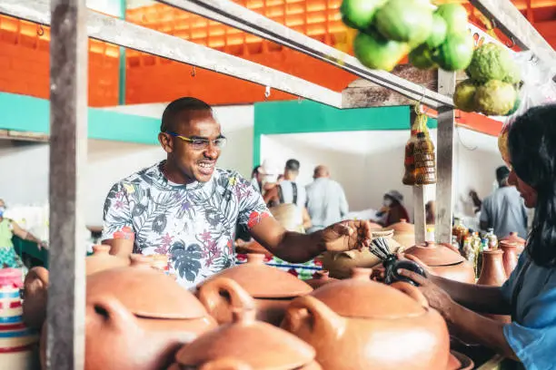smiling young market vendor behind his pottery stall showing his products to female client at public market in Bahia, Brazil
