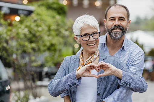 Cheerful mature couple is embracing on the street, they are looking at the camera and woman is making hear shape symbol with her hands