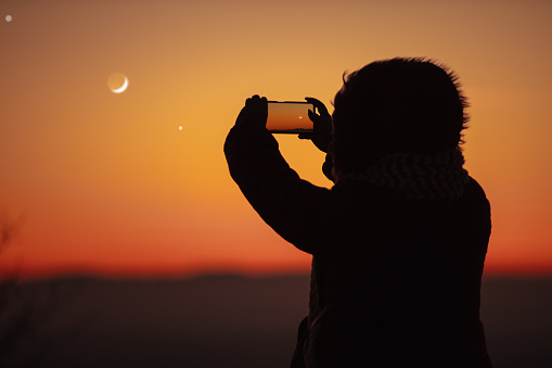 Woman taking photo with smartphone of evening sky with crescent Moon, stars and planets.