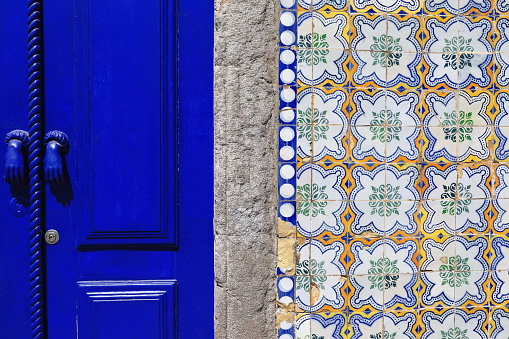 Facade detail-Neoclassical townhouse showing deep blue lacquered door leaf-plain stone door jamb-tiled wall of Portuguese azulejo in square pieces-floral and geometric motifs. Tavira-Algarve-Portugal.