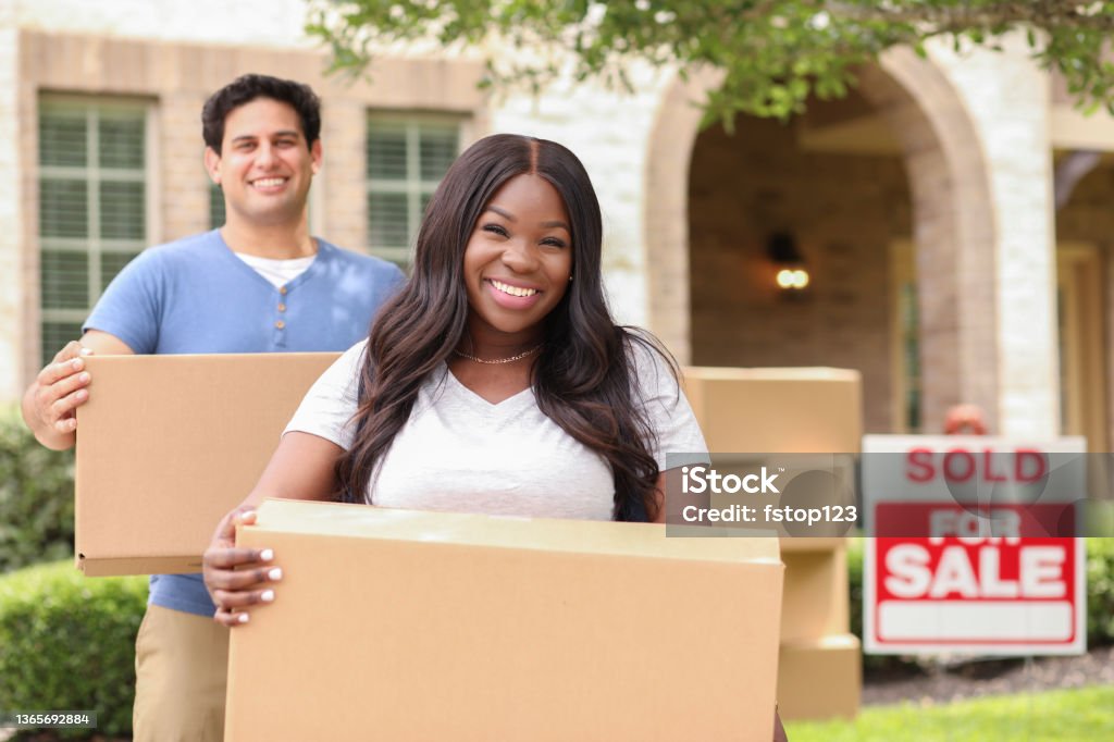 Excited young couple moves boxes into their new home. Mixed race family of African and Latin descent moving boxes into a new home.  Real estate sign and home in background.  Spring or summer season. Packing Stock Photo