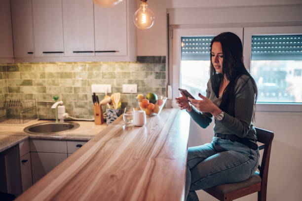 istock Middle aged woman using smartphone and drinking coffee in the kitchen at home 1365709735