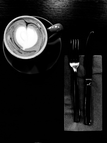 Vertical high contrast black and white high angle closeup photo of a mug of coffee latte with a heart foam art on a wooden table next to a knife, fork and paper serviette in a cafe.