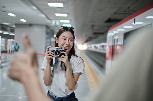 Young beautiful Asian female tourist taking portrait photo with film camera with friend, smile and enjoyment at train station platform, happy travel lifestyle by subway transport vacation trip.