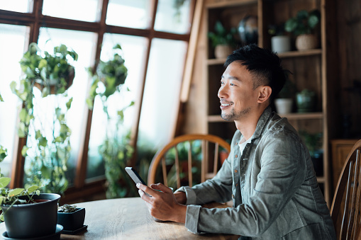 Portrait of smiling young Asian man with smartphone sitting at home by the window with green plants, relaxing and having a quiet moment