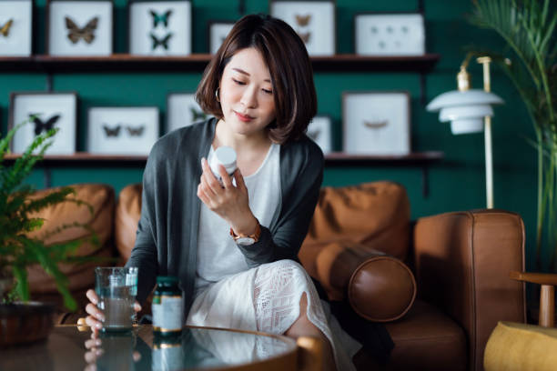 joven asiática tomando medicamentos con un vaso de agua en la mesa de café, leyendo la información en la etiqueta de su medicamento en casa. concepto de salud - drug label fotografías e imágenes de stock