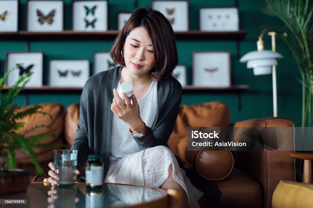 Young Asian woman taking medicines with a glass of water on the coffee table, reading the information on the label of her medication at home. Healthcare concept Nutritional Supplement Stock Photo