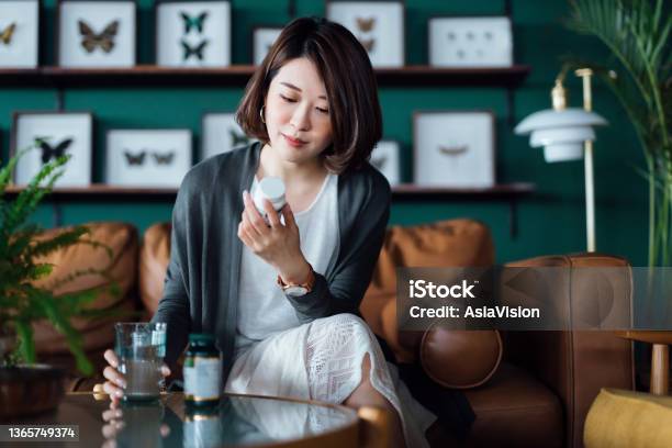 Young Asian Woman Taking Medicines With A Glass Of Water On The Coffee Table Reading The Information On The Label Of Her Medication At Home Healthcare Concept Stock Photo - Download Image Now