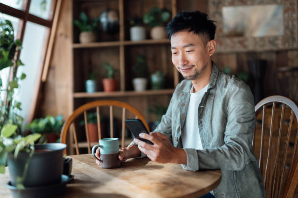 smiling young asian man using smartphone and having coffee in the balcony at home. the use of technology in everyday life - men asia asian culture asian ethnicity imagens e fotografias de stock