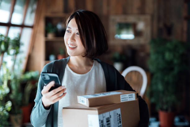 wanita muda asia yang tersenyum cantik dengan smartphone, menerima paket dengan layanan pengiriman rumah di rumah. belanja online, pembayaran mobile. pengalaman belanja yang menyenangkan - home delivery potret stok, foto, & gambar bebas royalti