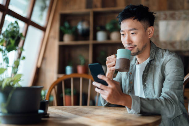 young asian man managing online banking with mobile app on smartphone, taking care of his money and finances while relaxing at home. banking with technology - smart phone text messaging mobile phone telephone imagens e fotografias de stock