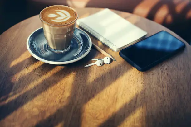 Close-up, latte coffee in old style glass and mobile phone, earphone, note book on wooden table with sunlight, vintage tone