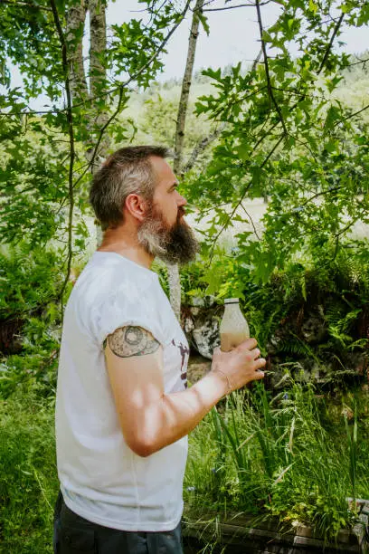 Picture of a tattooed man in white t-shirt drinking a healthy smoothie outside surrounded by green nature landscape in the french countryside.