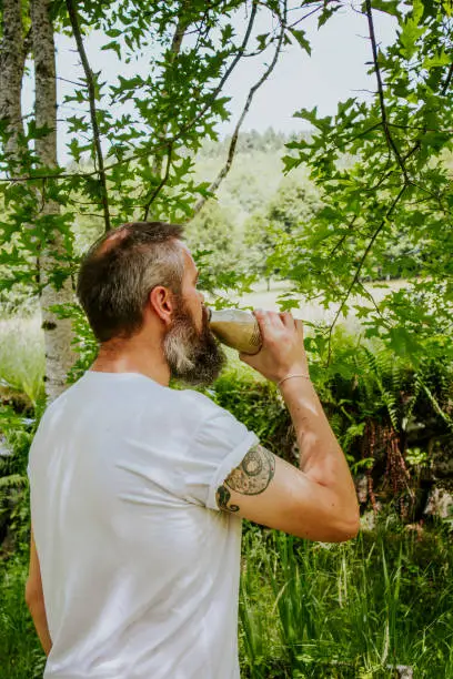 Picture of a tattooed man in white t-shirt drinking a healthy smoothie outside surrounded by green nature landscape in the french countryside.