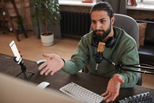 istock Horizontal medium portrait of modern young Middle Eastern man with beard on face sitting at desk speaking to followers during online stream 1365809657