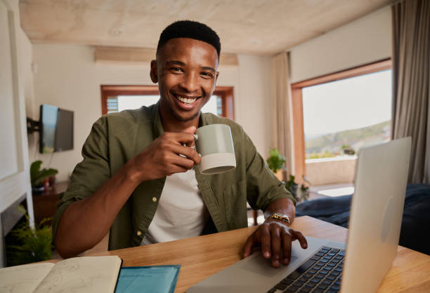 ritratto di giovane adulto maschio nero che tiene la tazza, sorridente alla macchina fotografica. lavorare da remoto in un appartamento moderno - ewan foto e immagini stock