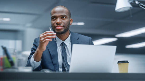 istock Office: Happy Successful Black Businessman Sitting at Desk Using Laptop Computer. African American Entrepreneur in Suit working with Stock Market Investing. Motion Blur Background. 1365824183