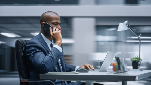 Modern Office: Productive Black Businessman Sitting at His Desk Working on a Computer, Answering Phone Call. African American Entrepreneur Wearing Suit working with Investment. Motion Blur Background