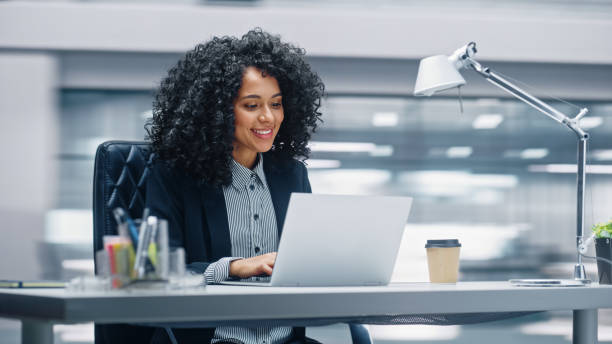modernes büro: schwarze geschäftsfrau sitzt an ihrem schreibtisch und arbeitet an einem laptop. lächeln erfolgreiche afroamerikanerin, die mit big data e-commerce arbeitet. bewegungsunschärfehintergrund - frau stock-fotos und bilder