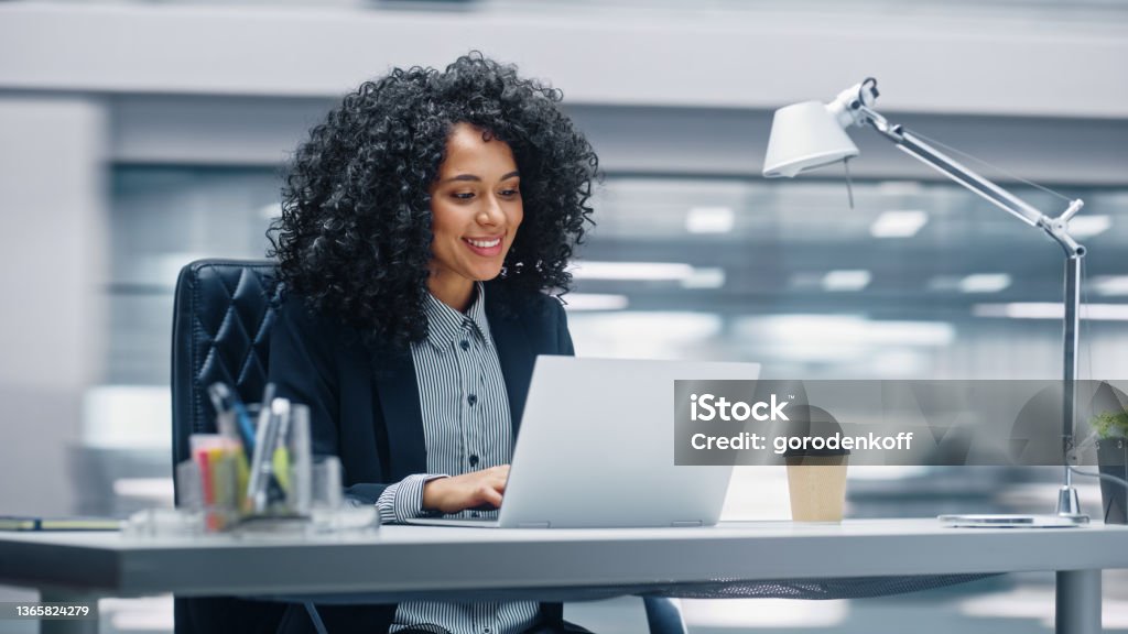 Modern Office: Black Businesswoman Sitting at Her Desk Working on a Laptop Computer. Smiling Successful African American Woman working with Big Data e-Commerce. Motion Blur Background Office Stock Photo