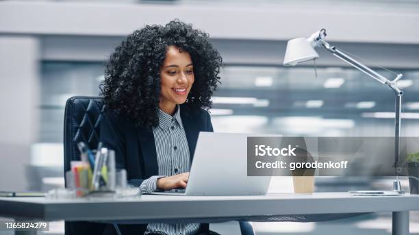 Modern Office Black Businesswoman Sitting At Her Desk Working On A Laptop Computer Smiling Successful African American Woman Working With Big Data Ecommerce Motion Blur Background Stock Photo - Download Image Now