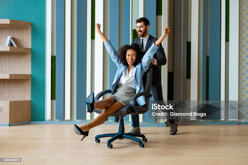 Happy and playful business people Two young cheerful business people in business wear having fun while racing on office chairs and smiling - Colleagues celebrating a successful deal at work, happy and joyful moments in the office Office Chair Stock Photo