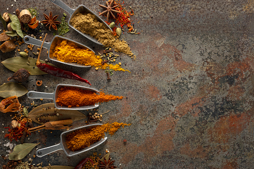 Many colorful, organic, dried, vibrant Indian food, ingredient spices in small aluminum metal spice scoops are arranged in a line on an old, weathered, rusted metal plate background. Taken directly above. Good copy space to the right of the image.