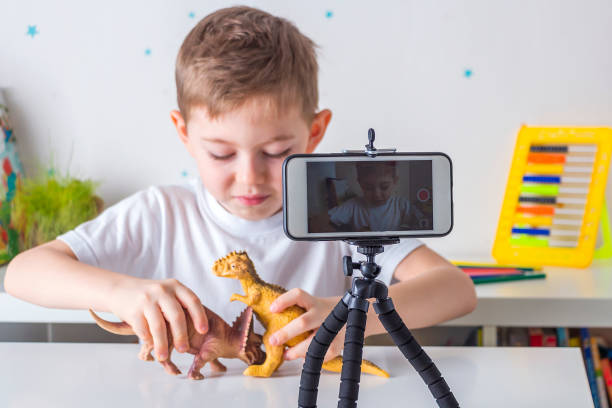 Happy little boy (preschooler) blogger recording live video streaming on smartphone. Online unboxing of dinosaures toys and plays it to followers. Blogging concept, backlight, selective focus feliz niño pequeño (preescolar) blogger grabando transmisión de video en vivo en el teléfono inteligente. desempaquetado en línea de juguetes de dinosaurios y se lo juega a los seguidores. concepto de blogging, retroiluminación, enfoque selectivo.