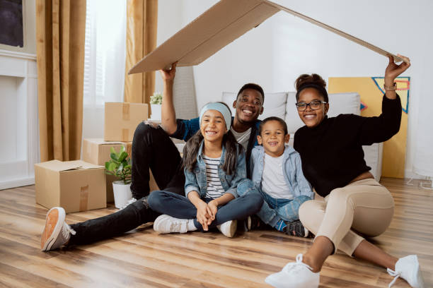 family complete, parents with two children sitting on the floor relaxing after moving to a new apartment, holding over their heads a piece of cardboard left from boxes as a roof, smiling at the camera - family large american culture fun imagens e fotografias de stock