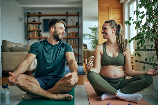 Pregnant woman and her husband sitting in lotus pose at home and looking at each other