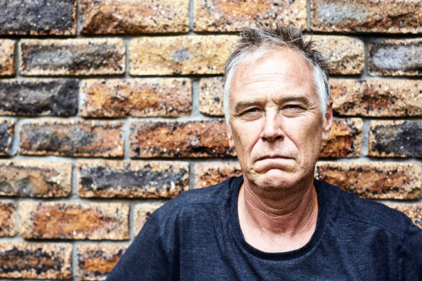 Mature man stares grimly directly at the camera, looking serious and displeased Sad, stern man in his 50s looks at the camera against a brick wall. squint eyes stock pictures, royalty-free photos & images
