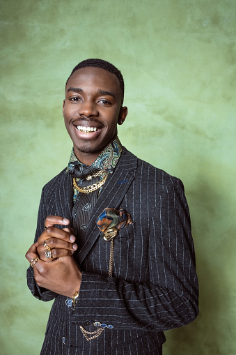 Fashion portrait of handsome young man wearing suit jacket, printed shirt, gold jewelry, smiling at camera. Studio shot on green background.