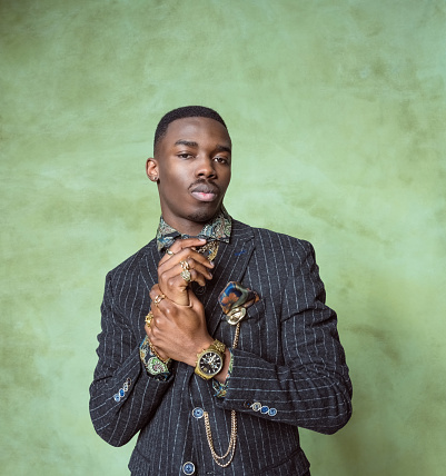 Fashion portrait of handsome young man wearing jacket, printed shirt, gold jewelry, looking at camera. Studio shot on green background.
