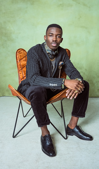 Fashion portrait of handsome young man wearing jacket, printed shirt, gold jewelry, sitting on leather armchair. Studio shot on green background.
