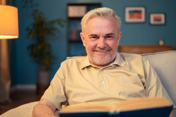 istock Portrait of a smiling, contented elderly man sitting on a couch looking into a camera. Elderly man holding a book, an old book. Grey haired guy with stubble contentedly sitting in living room on couch 1365842368