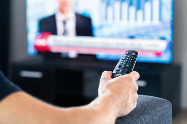 istock Man watching tv news program and sitting on couch home. Television remote control in hand. Politics, business or finance network channel. Online live broadcast in monitor screen. 1365843280