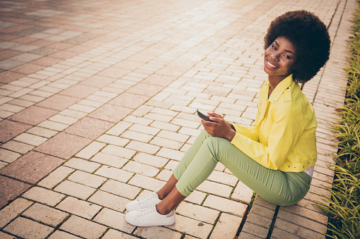 Photo portrait of woman holding phone in two hands sitting on curb outdoors.