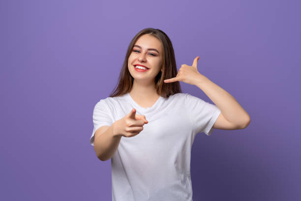 young beautiful brunette girl wearing white t shirt standing over purple background smiling doing talking on the telephone gesture and pointing to you. call me - american sign language imagens e fotografias de stock