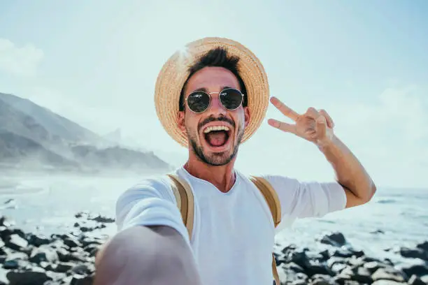 Photo of Happy handsome man taking selfie outside - Smiling guy having fun on the beach - Mobile, travel and people lifestyle concept