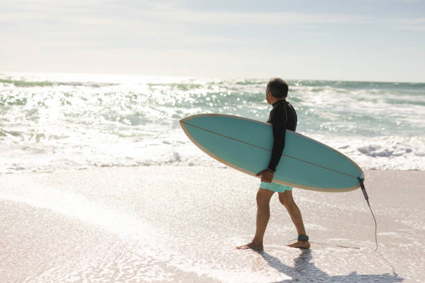 istock Biracial senior man carrying surfboard wading on shore looking at waves splashing during sunny day 1365855809