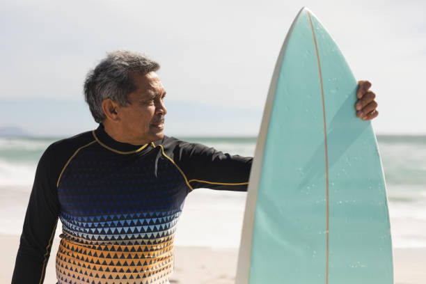 istock Biracial senior man looking away while standing with surfboard at beach on sunny day 1365858053