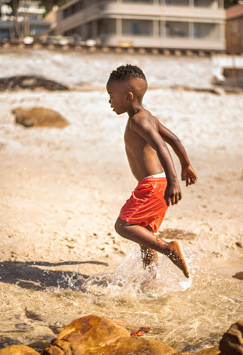 Cute little African boy in swim shorts splashing in the sea during a trip to the beach in summer