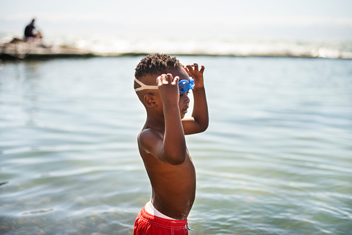 Adorable little African boy wearing swim shorts and goggles standing in the sea during a trip to the beach in summer